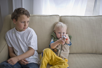 Young toddler aiming a toy gun while seated next to his older brother on a sofa in the living room.