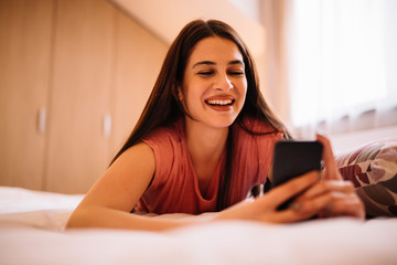 Beautiful smiling girl with smartphone is lying on bed at home