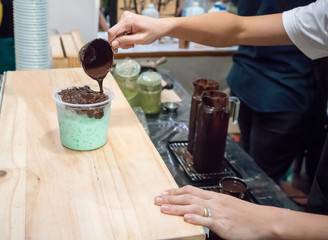 Woman hand holding silver cup adding chocolate syrup on top of green milk tea in plastic cup