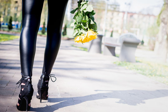 Legs view of a woman walking with flowers. Woman wear high heels shoes. The girl in black pants moves around park outside. Beuty and fashion concept.