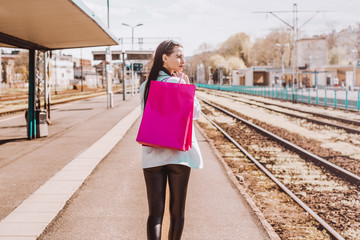 A woman is waiting for the train to look out for a train among shopping bags. The woman returns home from shopping.