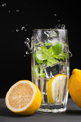 The concept of holidays and parties, food and drinks. Cold alcoholic cocktail mojito, lemonade with lemon, mint and ice in a glass and a bottle on a black background. Vertical frame, with spray