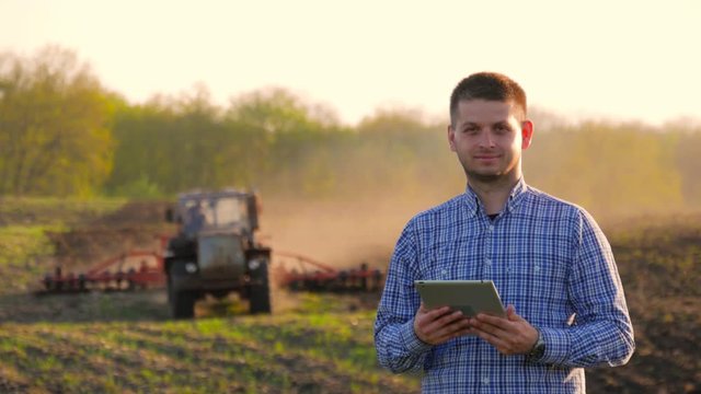 Young farmer holding tablet in front of tractor in field. Farmer controls the work in the field over it's tablet. Seasonal agricultural workers.