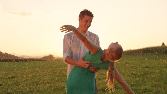 SLOW MOTION, CLOSE UP: Cheerful young man holds carefree blonde woman tightly as she leans back in his arms while they dance in the refreshing summer rain. Lovely Caucasian couple dancing outdoors.
