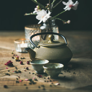 Traditional Asian tea ceremony arrangement. Golden iron teapot, cups, candles and almond blossom flowers over vintage wooden table background, selective focus, square crop