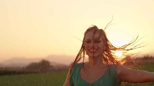 SLOW MOTION, PORTRAIT, LENS FLARE: Smiling girl gets drenched while she dances on a beautiful evening. Caucasian dancer having fun twirling in the rain and flipping her wet hair over her shoulder.