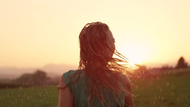 SLOW MOTION CLOSE UP LENS FLARE: Joyful blonde girl enjoys her evening in the countryside by dancing in the rain. Stunning golden sun rays shine on playful woman spinning and enjoying a spring shower.