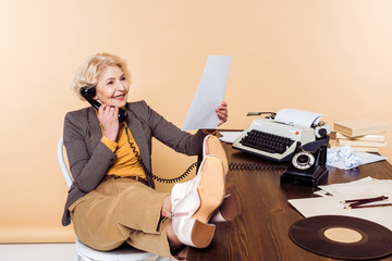 smiling senior woman with legs on table talking on rotary phone and looking at pape