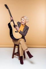 Fototapeta premium stylish senior woman sitting on chair and playing on acoustic guitar