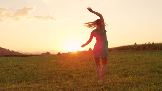 SLOW MOTION, LENS FLARE: Relaxed young Caucasian woman dances freely in a beautiful meadow at sunrise. Attractive blonde haired girl spinning in rain on a tranquil evening in the idyllic countryside.