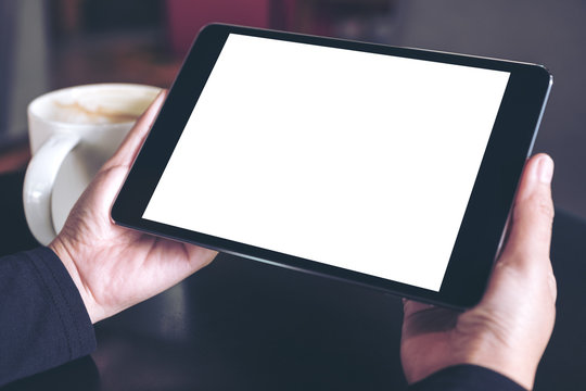 Top view mockup image of hands holding black tablet pc with white blank screen and coffee cup on table background