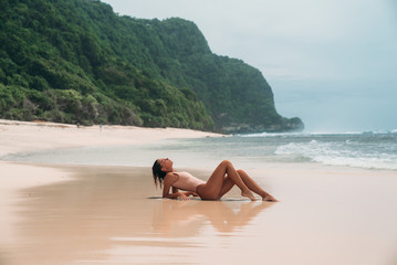Fototapeta na wymiar gorgeous young girl in a beige bikini lies on the beach, sunbathing near the ocean. A model with a sexy body and a sports figure in the clear sea water on the island. The concept of the weekend on