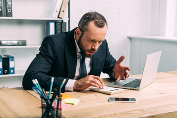 shocked and stressed businessman with calculator at workplace with laptop in office