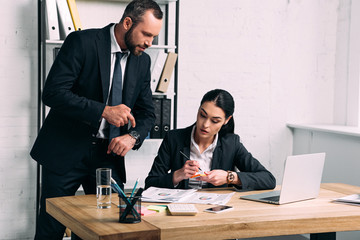 businessman pointing at watch near concentrated businesswoman at workplace with laptop in office