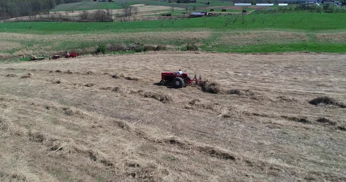 Side aerial view of tractor going through a field of switchgrass with a star wheel rake raking the grass.