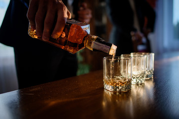 Man pours whisky in the glasses standing before a wooden table