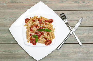 Penne pasta with vegetables and parmesan cheese on a white square plate on a wooden table. Top view