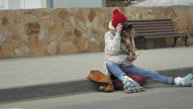 Young beautiful woman in red hat wearing sporty warm clothes and rollers, sitting on the asphalt road and taking pictures on a vintage camera