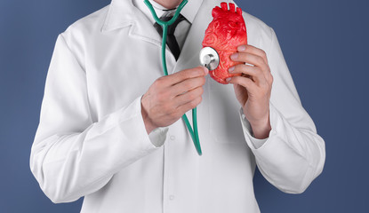 Doctor holding model of heart and stethoscope on color background. Prevent heart attack