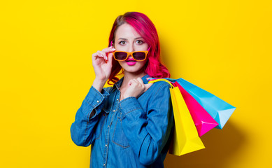 Young pink hair girl in sunglasses and blue shirt holding a colored shopping bags. Portrait on isolated yellow background