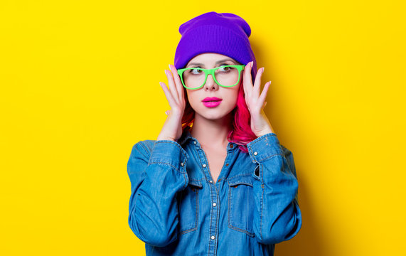 Young pink hair girl in blue shirt, purple hat and green glasses. Portrait isolated on yellow background
