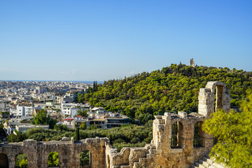View of Athens cityscape and the Philopappu Monument through ancient stone theatre seeing lowrise white buildings architecture, mountain, trees and clear blue sky background