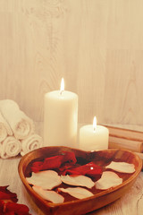 Wooden bowl in the shape of heart with rose petals and nearby burning candles, preparation for spa procedures