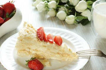 A piece of Napoleon cake on a plate and a bowl of strawberries, next to a glass of milk and a bouquet
