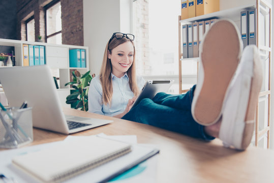 Portrait of charming, pretty, attractive, cheerful woman with glasses on head put her legs on the table, searching, watching, looking something on tablet, wearing jeans, pants