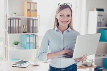 Portrait of charming, nice, positive woman with glasses on head having laptop in hands looking at camera standing in work place, station