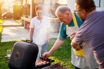 Elderly woman is observing and laughing while her husband is carefully rotating meat and vegetables on the grill.