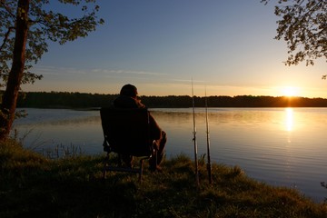 Fisherman sitting on the chair and fishing on the shore of lake during sunrise
