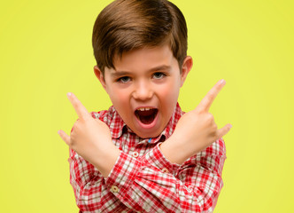 Handsome toddler child with green eyes making rock symbol with hands, shouting and celebrating over yellow background