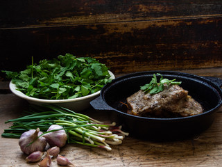 Tasty juicy meat in a frying pan. Garlic and leaves of young garlic are sliced on a board. Warehouse on a wooden background. Spring greens.