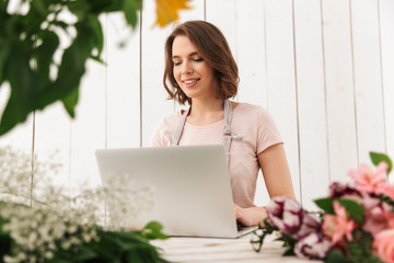 Cheerful florist lady standing with flowers using laptop computer.