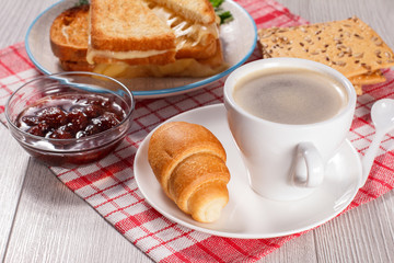 Cup of coffee and croissant on saucer, glass bowl with strawberry jam, cookies with whole grains of sunflower seeds and toasted slices of bread with cheese