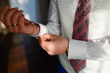 The man fixes the cuffs on the white shirt and stylish vest and blue pants with expensive gold accessories, watches, rings and rings. The businessman is preparing to meet in an expensive interior.