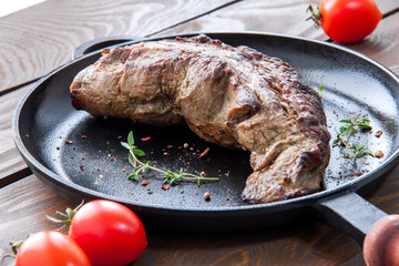 roast beef fillet on a black iron frying pan with seasonings and herbs and a twig branch on a wooden table with red ripe tomatoes