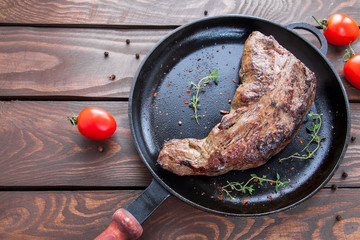 fried beef meat with herbs and spices on a black iron frying pan on a wooden table with red ripe cherry tomatoes