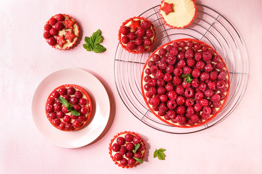 Variety of red raspberry shortbread tarts and tartlets with lemon custard and glazed fresh raspberries served on cooling rack over pink pastel background. Top view, space.
