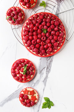 Variety of red raspberry shortbread tarts and tartlets with lemon custard and glazed fresh raspberries served on cooling rack over white marble texture background. Top view, space.