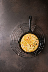 Spanish potato omelette tortilla with bacon served in cast-iron pan on cooling rack over dark brown texture background. Top view, space.