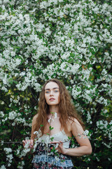 A beautiful girl in a dress stands next to a blooming cherry bush.