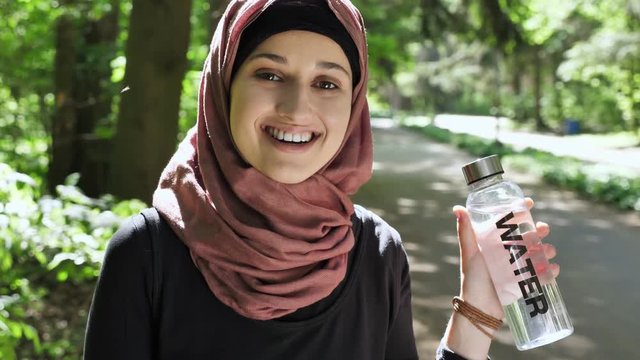 Portrait of a cute young girl in a hijab with a bottle of water in her hands, smiling, looking at the camera, park in the background. 50 fps