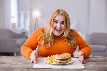 Favourite food. Cheerful overweight woman sitting at the table and eating fries and sandwiches