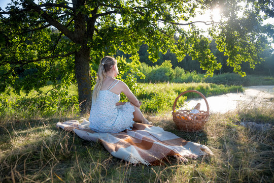 Rear view image of young woman in summer dress sitting under big tree and looking at sunset