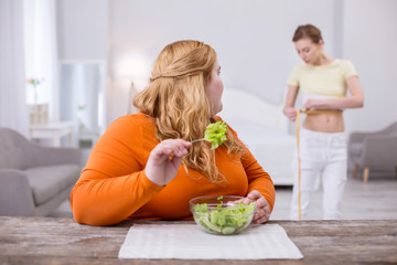 Envious. Young overweight woman eating a salad and looking at her slim friend