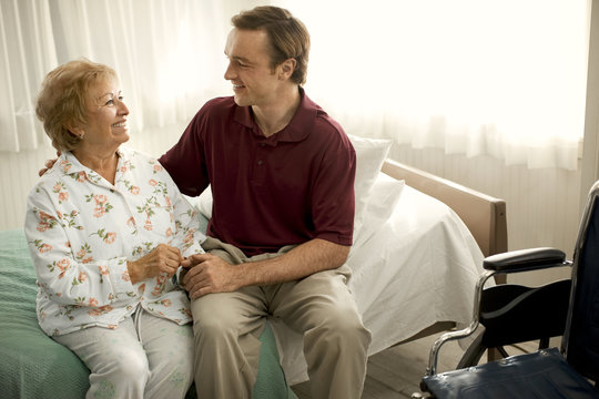 Smiling senior woman speaking with a male nurse on the edge of a bed while holding his hand.