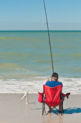 Back view of a senior, white, male siting and relaxing on a tropical beach, surf fishing with a caught fish on a long fishing pole on a sunny, winter day, on Gulf of Mexico