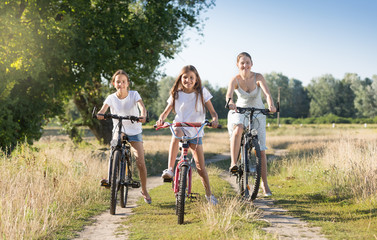 Two smiling girls riding bicycles with mother on countryside road